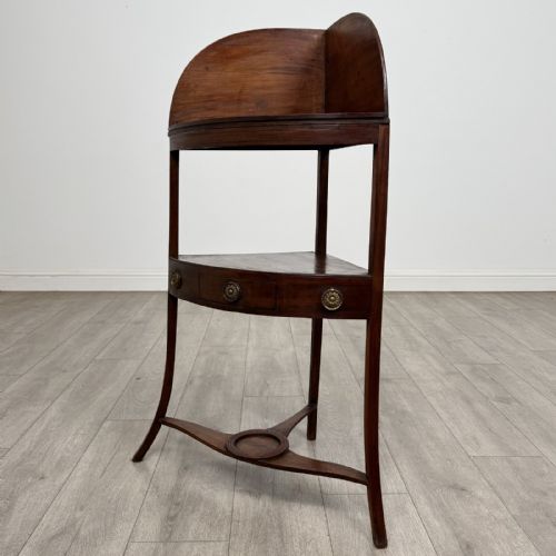 fig 1. A Georgian style mahogany corner washstand with a high curved splashback gallery, central drawer tier with brass rosette handles, and splayed legs joined by a circular lower stretcher.