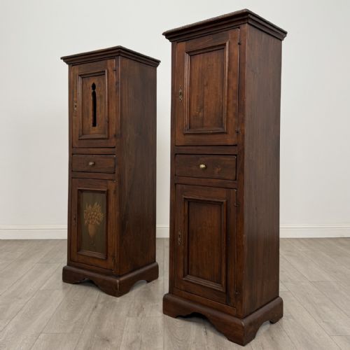 fig 2. A pair of tall dark brown hardwood cupboards featuring a central drawer and panelled doors, with one lower panel decorated with a painted urn motif.