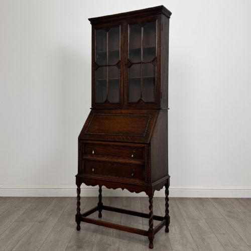 fig 2. A dark oak Jacobean style bureau bookcase featuring twin glazed cabinet doors above a slant-front writing desk with two lower drawers and barley twist legs.