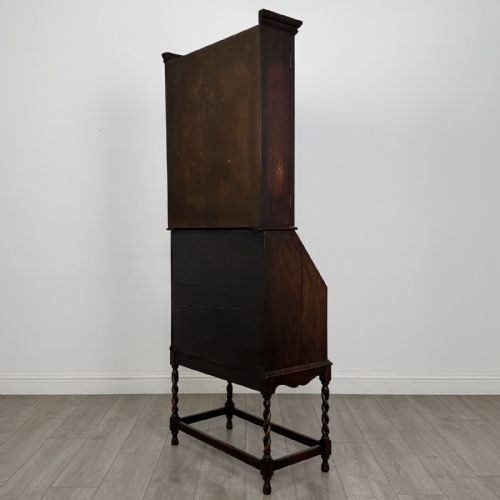 fig 6. A dark oak Jacobean style bureau bookcase featuring twin glazed cabinet doors above a slant-front writing desk with two lower drawers and barley twist legs.