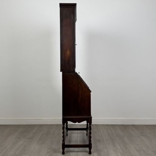 fig 7. A dark oak Jacobean style bureau bookcase featuring twin glazed cabinet doors above a slant-front writing desk with two lower drawers and barley twist legs.