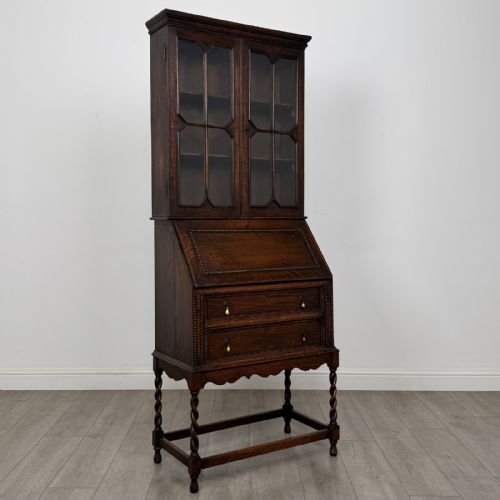 fig 8. A dark oak Jacobean style bureau bookcase featuring twin glazed cabinet doors above a slant-front writing desk with two lower drawers and barley twist legs.