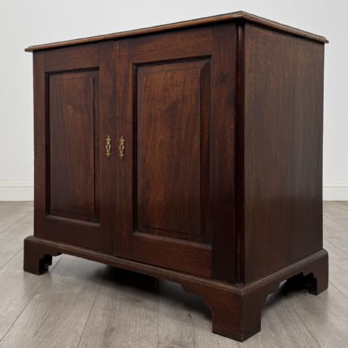 fig 1. An 18th-century dark mahogany low cupboard featuring twin panelled doors with brass keyhole escutcheons resting on bracket feet.