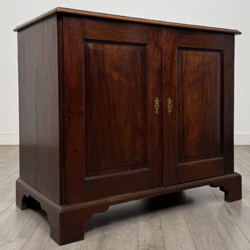 fig 7. An 18th-century dark mahogany low cupboard featuring twin panelled doors with brass keyhole escutcheons resting on bracket feet.