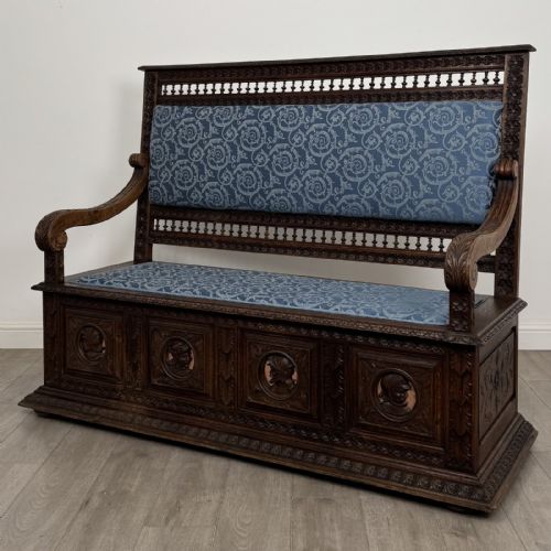 fig 1. A dark carved oak hall bench with patterned blue upholstered seat and backrest cushions, spindled galleries, and relief-carved profile medallions across the base.
