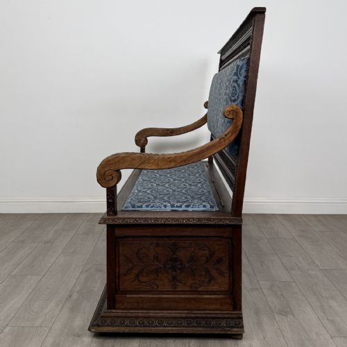 fig 2. A dark carved oak hall bench with patterned blue upholstered seat and backrest cushions, spindled galleries, and relief-carved profile medallions across the base.