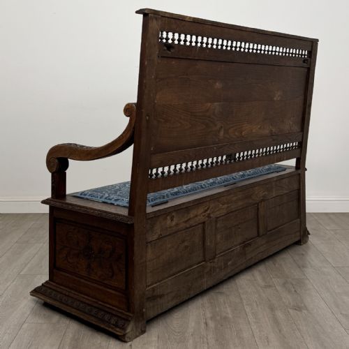 fig 3. A dark carved oak hall bench with patterned blue upholstered seat and backrest cushions, spindled galleries, and relief-carved profile medallions across the base.