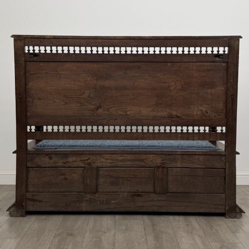 fig 4. A dark carved oak hall bench with patterned blue upholstered seat and backrest cushions, spindled galleries, and relief-carved profile medallions across the base.