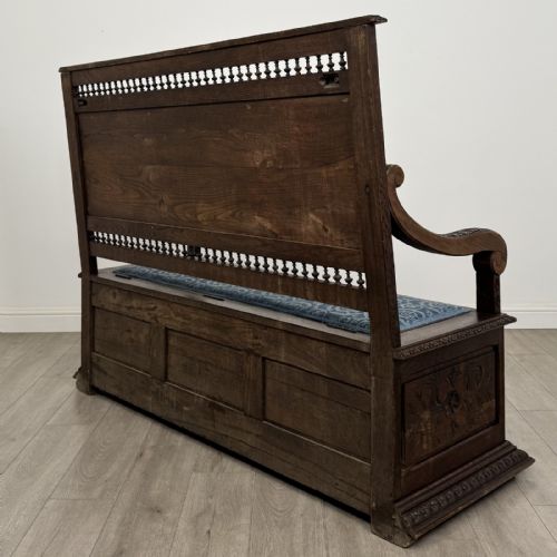 fig 5. A dark carved oak hall bench with patterned blue upholstered seat and backrest cushions, spindled galleries, and relief-carved profile medallions across the base.