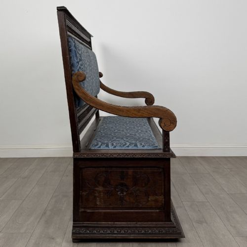 fig 6. A dark carved oak hall bench with patterned blue upholstered seat and backrest cushions, spindled galleries, and relief-carved profile medallions across the base.