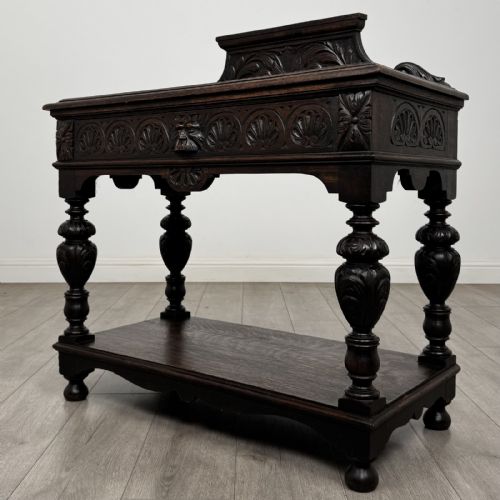 fig 1. A dark carved oak side table featuring a Green Man mask on the front frieze, turned legs, a low undertier, and a raised carved gallery back.