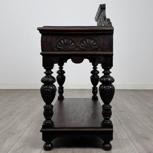 fig 2. A dark carved oak side table featuring a Green Man mask on the front frieze, turned legs, a low undertier, and a raised carved gallery back.
