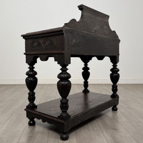 fig 3. A dark carved oak side table featuring a Green Man mask on the front frieze, turned legs, a low undertier, and a raised carved gallery back.
