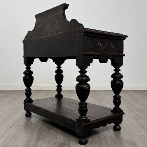 fig 5. A dark carved oak side table featuring a Green Man mask on the front frieze, turned legs, a low undertier, and a raised carved gallery back.