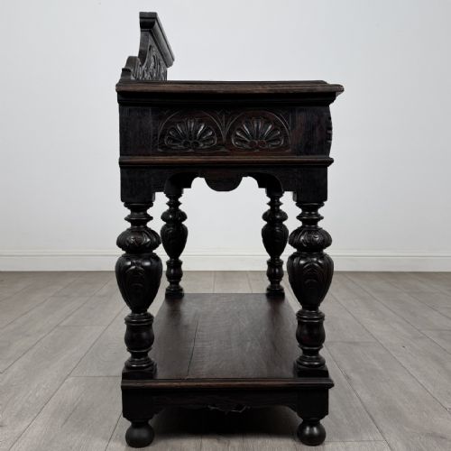 fig 6. A dark carved oak side table featuring a Green Man mask on the front frieze, turned legs, a low undertier, and a raised carved gallery back.
