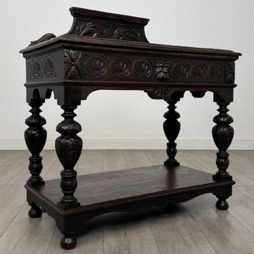 fig 7. A dark carved oak side table featuring a Green Man mask on the front frieze, turned legs, a low undertier, and a raised carved gallery back.