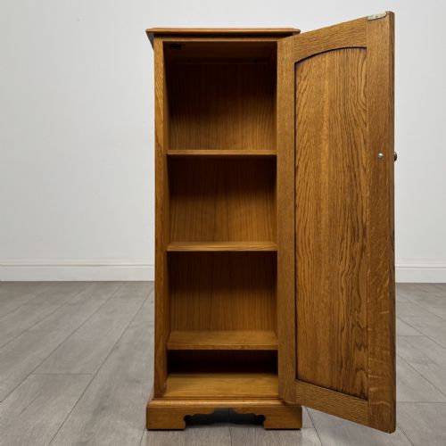 fig 1. A tall, narrow warm oak cupboard featuring a single arched fielded panel door with three brass H-hinges and a small brass door knob.