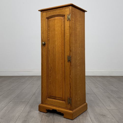fig 2. A tall, narrow warm oak cupboard featuring a single arched fielded panel door with three brass H-hinges and a small brass door knob.
