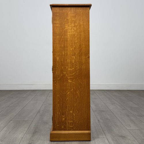 fig 3. A tall, narrow warm oak cupboard featuring a single arched fielded panel door with three brass H-hinges and a small brass door knob.