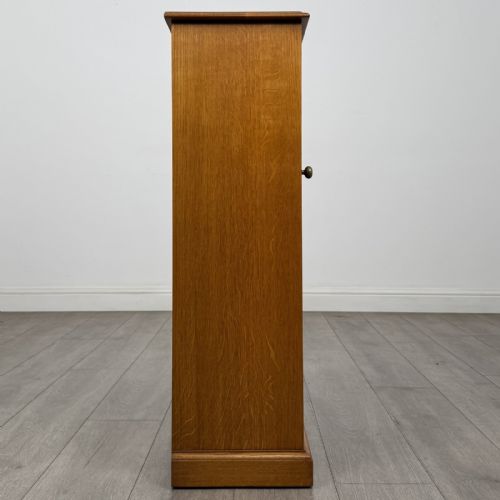 fig 7. A tall, narrow warm oak cupboard featuring a single arched fielded panel door with three brass H-hinges and a small brass door knob.
