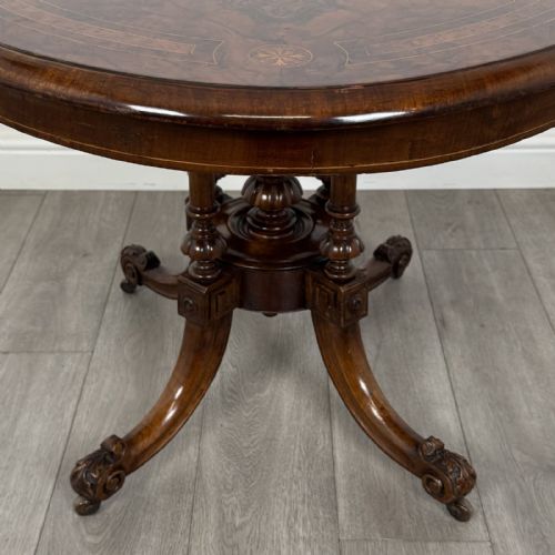 fig 5. A Victorian burr walnut tilt-top loo table featuring an oval top supported by four turned columns and carved quadform legs with scroll feet.