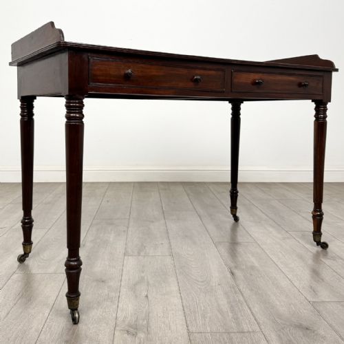 fig 7. A dark mahogany Georgian writing desk featuring two frieze drawers with wooden knobs, turned legs on brass castors, and a raised gallery edge.