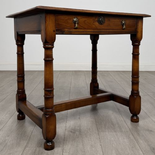 fig 7. A warm brown fruitwood side table featuring a single drawer with twin brass drop handles, turned legs, and an H-stretcher.