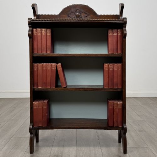 fig 1. An Arts and Crafts dark oak open bookcase featuring three shelves, a light blue painted back panel, and a carved cresting gallery.