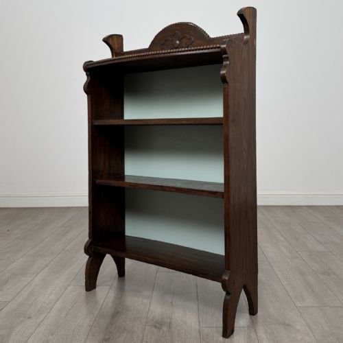 fig 2. An Arts and Crafts dark oak open bookcase featuring three shelves, a light blue painted back panel, and a carved cresting gallery.
