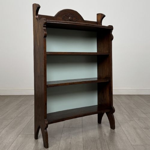 fig 8. An Arts and Crafts dark oak open bookcase featuring three shelves, a light blue painted back panel, and a carved cresting gallery.