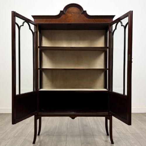 fig 1. An Edwardian mahogany display cabinet featuring two astragal glazed doors, a shaped arched cresting, and lower veneered panels supported on slender splayed legs.