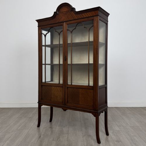 fig 2. An Edwardian mahogany display cabinet featuring two astragal glazed doors, a shaped arched cresting, and lower veneered panels supported on slender splayed legs.