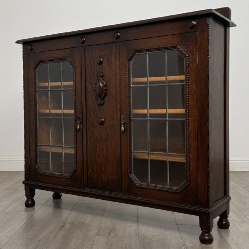 fig 1. A dark oak Jacobean style display cabinet featuring twin leaded glass doors, central carved bosses, internal shelving, and turned feet.