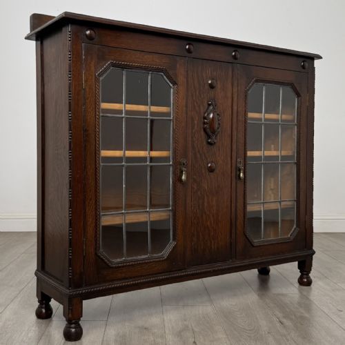fig 7. A dark oak Jacobean style display cabinet featuring twin leaded glass doors, central carved bosses, internal shelving, and turned feet.