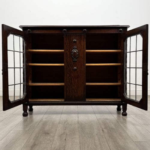 fig 8. A dark oak Jacobean style display cabinet featuring twin leaded glass doors, central carved bosses, internal shelving, and turned feet.