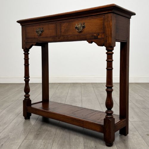 fig 1. A narrow dark wood console table featuring two drawers with brass handles, turned front legs, and a lower potboard shelf.