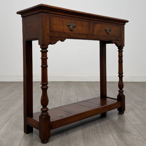 fig 7. A narrow dark wood console table featuring two drawers with brass handles, turned front legs, and a lower potboard shelf.