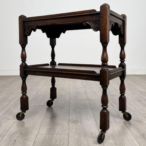 fig 1. A dark brown oak two-tier tea trolley featuring turned supports, carved corner spandrels, and metal castors on the feet.