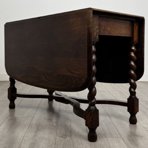 fig 1. A dark brown oak drop-leaf dining table with folded side leaves, barley twist legs, and a shaped cross-stretcher base.
