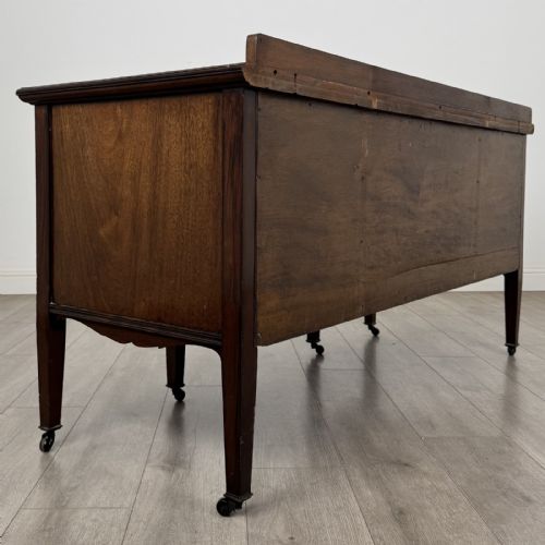 fig 3. An Edwardian mahogany sideboard featuring detailed foliate marquetry inlay, seven drawers with brass handles, an arched central apron, and tapered legs with castors.