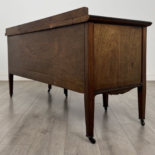 fig 5. An Edwardian mahogany sideboard featuring detailed foliate marquetry inlay, seven drawers with brass handles, an arched central apron, and tapered legs with castors.
