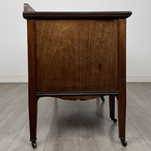 fig 6. An Edwardian mahogany sideboard featuring detailed foliate marquetry inlay, seven drawers with brass handles, an arched central apron, and tapered legs with castors.