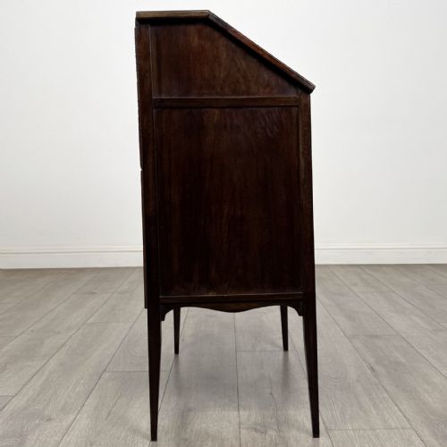 fig 6. An Edwardian dark mahogany bureau featuring a slant-front writing lid, a single drawer with brass drop handles, open lower compartment with arches, and tapered legs.
