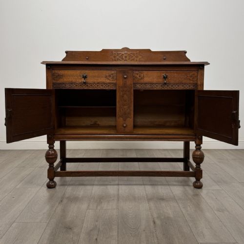 fig 1. A Jacobean style oak sideboard featuring two drawers, carved door panels, metal drop handles, and turned cup and cover legs connected by low stretchers.