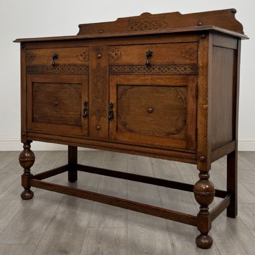 fig 2. A Jacobean style oak sideboard featuring two drawers, carved door panels, metal drop handles, and turned cup and cover legs connected by low stretchers.