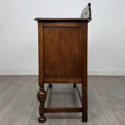 fig 3. A Jacobean style oak sideboard featuring two drawers, carved door panels, metal drop handles, and turned cup and cover legs connected by low stretchers.