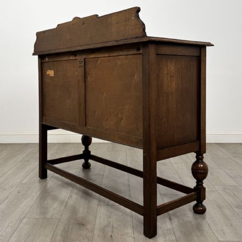 fig 6. A Jacobean style oak sideboard featuring two drawers, carved door panels, metal drop handles, and turned cup and cover legs connected by low stretchers.