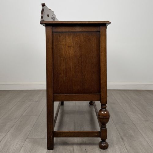 fig 7. A Jacobean style oak sideboard featuring two drawers, carved door panels, metal drop handles, and turned cup and cover legs connected by low stretchers.