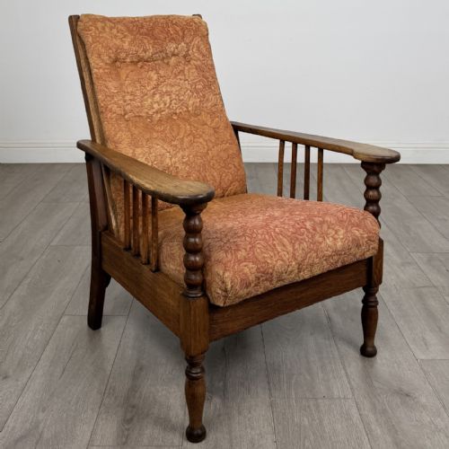 fig 7. An Arts and Crafts oak reclining armchair with wide armrests, turned front legs, vertical side spindles, and patterned terracotta upholstered cushions.