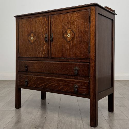 fig 1. A dark oak Arts and Crafts cupboard featuring double panelled doors with floral marquetry inlays above two wide drawers, finished with dark metal Celtic knot handles.